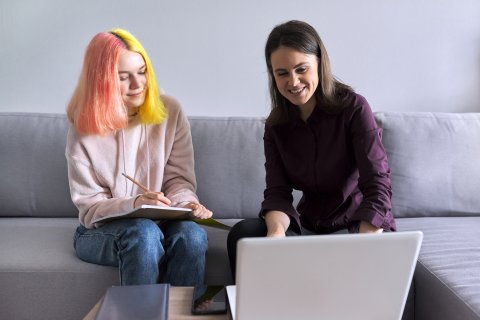 a young adult sitting on a couch with a woman looking at a laptop 