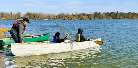 Two young adults are sitting in a canoe on a lake that is being held by an adult