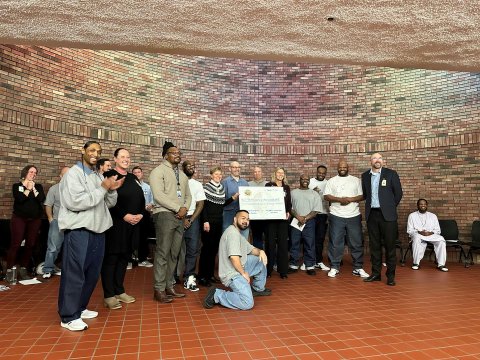 A group of diverse individuals standing together in front of a brick wall posing for a photo with two women in the center holding a giant check