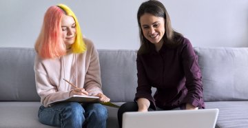 a young adult sitting on a couch with a woman looking at a laptop 