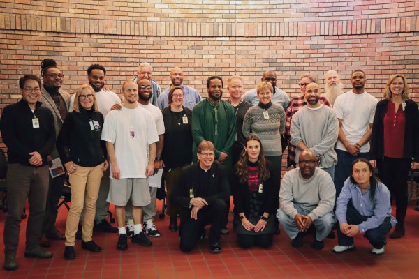 A diverse group of individuals facing the camera in front of a brick wall some standing while others are kneeling in front smiling