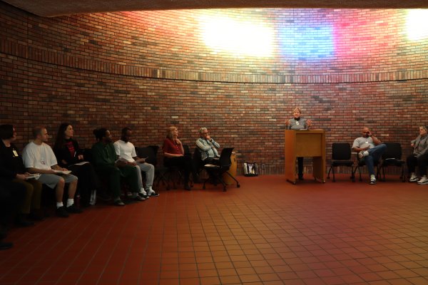 Diverse individuals sitting in chairs lined up against a brick wall paying attention to a woman speaking at a podium 