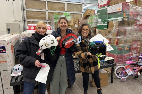 Three women in a warehouse setting holding up different styles of bicycle helmets and smiling 