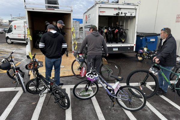 Three men facing away from the camera load bikes onto a trailer parked outside near a building